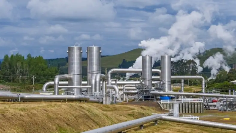 Industrial facility with insulated silver pipework and vertical tanks releasing steam against a green hillside